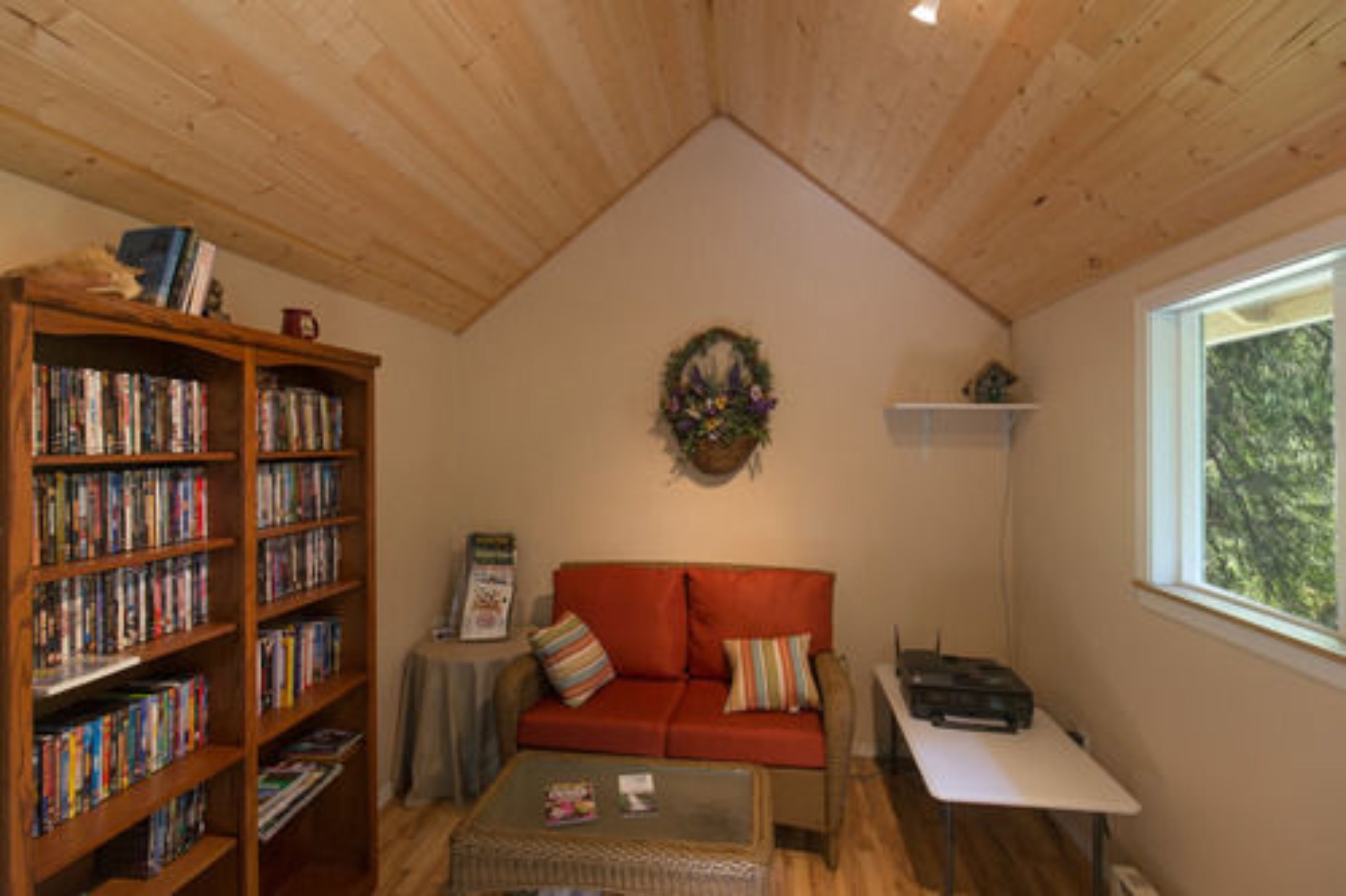 Een knus interieur met een houten plafond, een rode bank, een boekenkast, een kleine tafel en een raam.