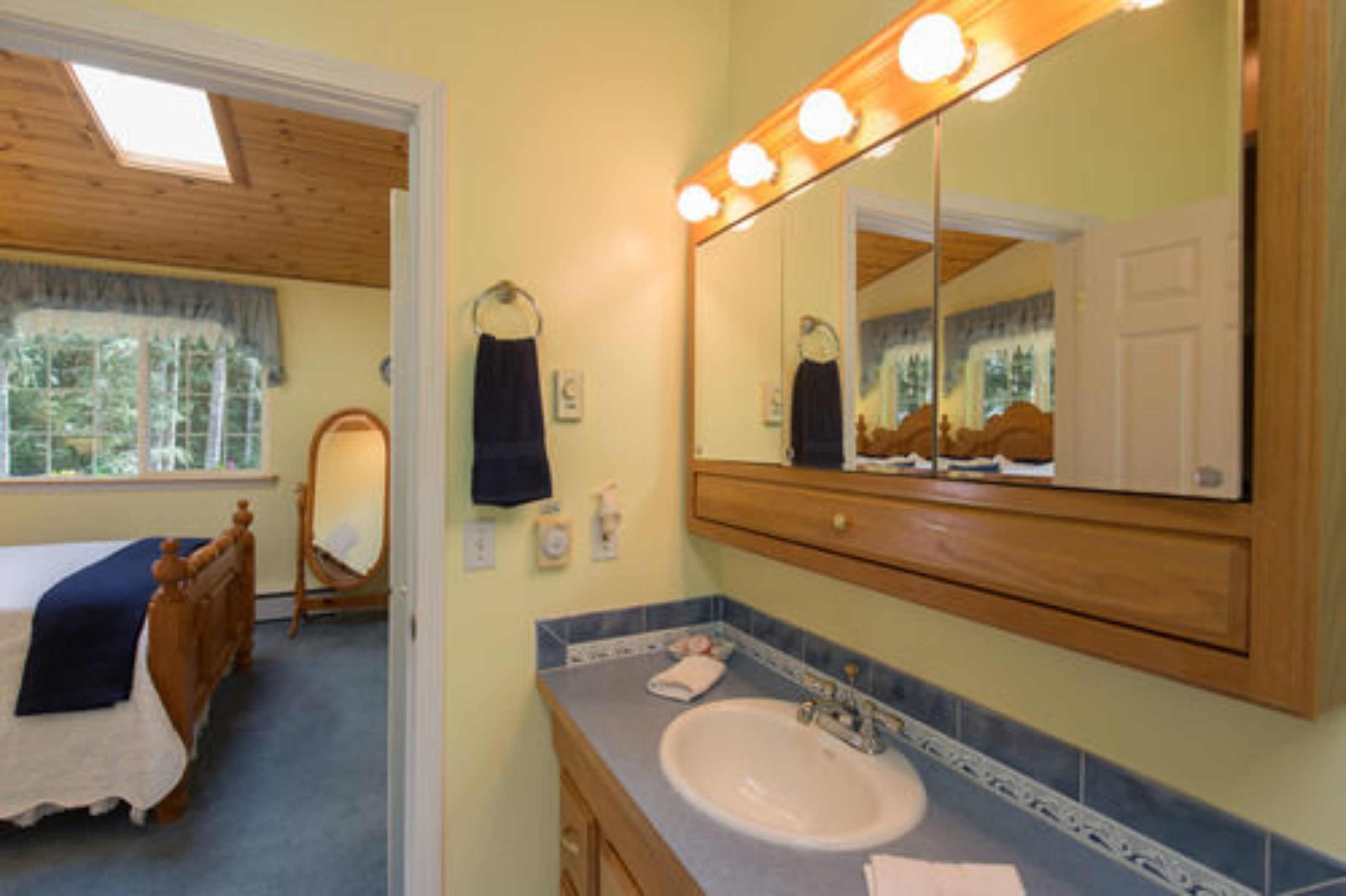 A bedroom with a bed, a wooden headboard, a nightstand, a mirror with lights, a window with curtains, and a bathroom with a sink and mirror.