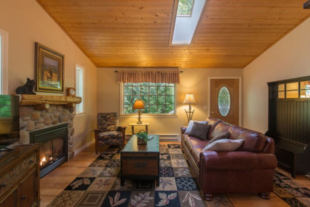 A cozy living room with a fireplace, leather sofa, patterned rug, and a large window with a view of greenery.