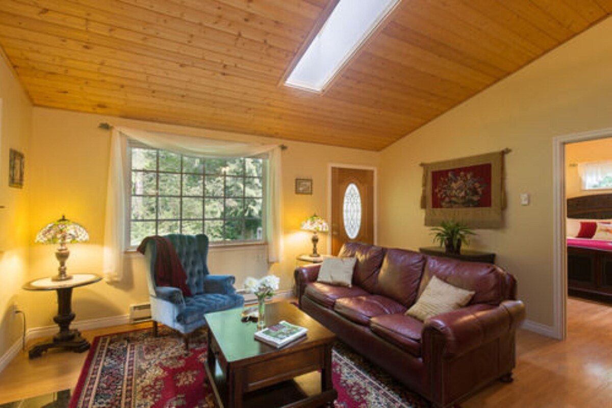 A cozy living room with a wooden ceiling, a large window, a leather sofa, a blue armchair, and a small table with a lamp.