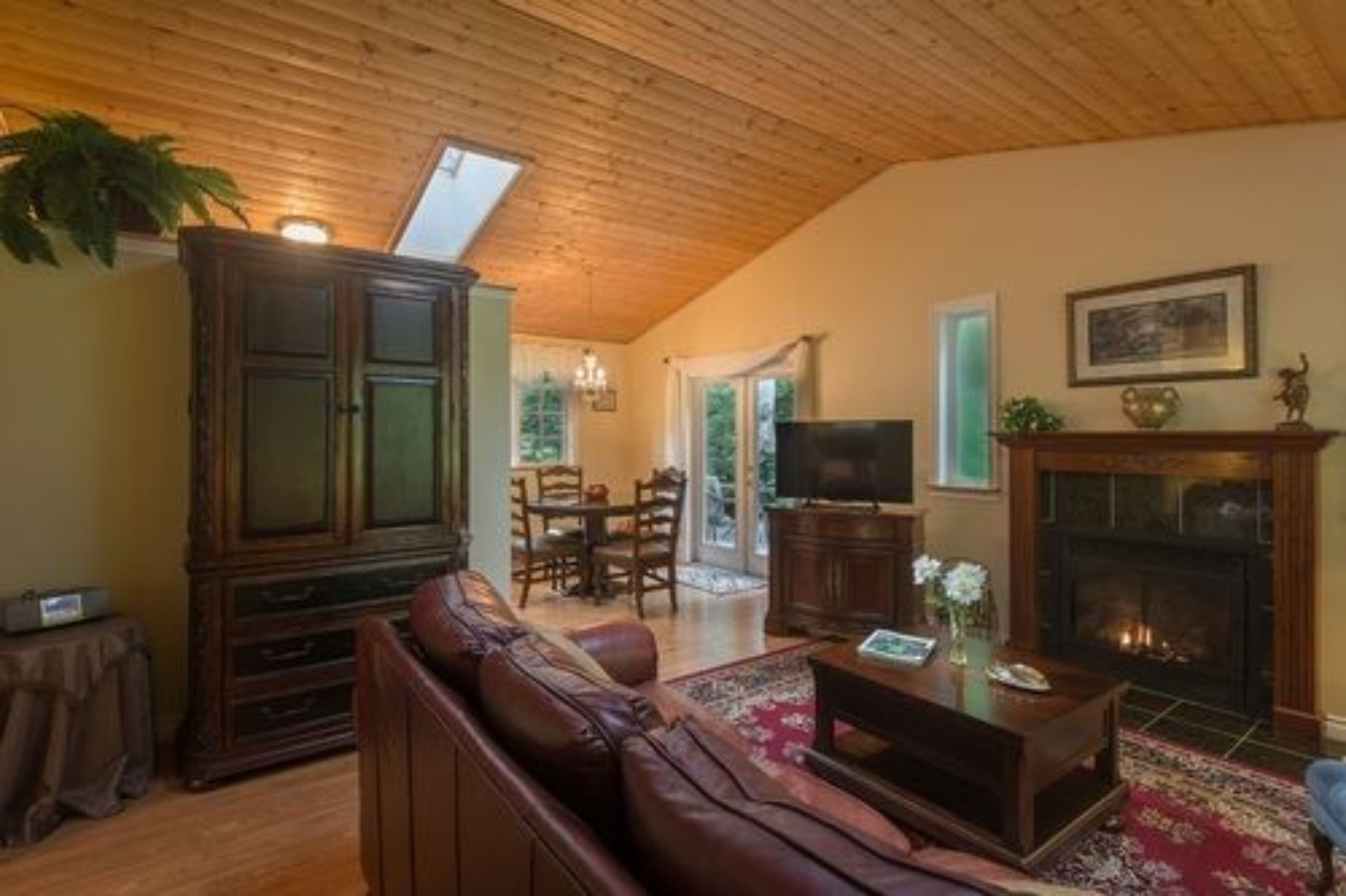 A cozy living room with a wooden ceiling, a fireplace, a television, and a dining area with chairs and a table.