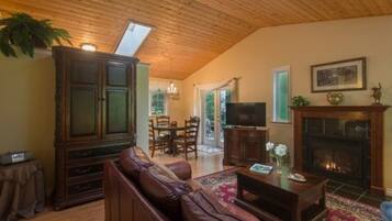 A cozy living room with a wooden ceiling, a fireplace, a television, and a dining area with chairs and a table.