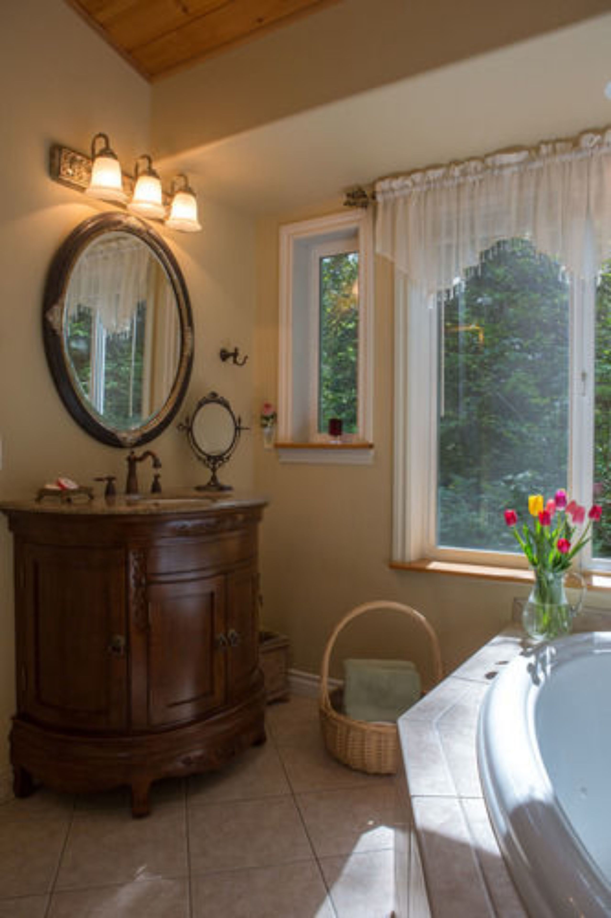 Un baño con tocador de madera, un espejo grande, una ventana con vistas a follaje y un jarrón con flores.