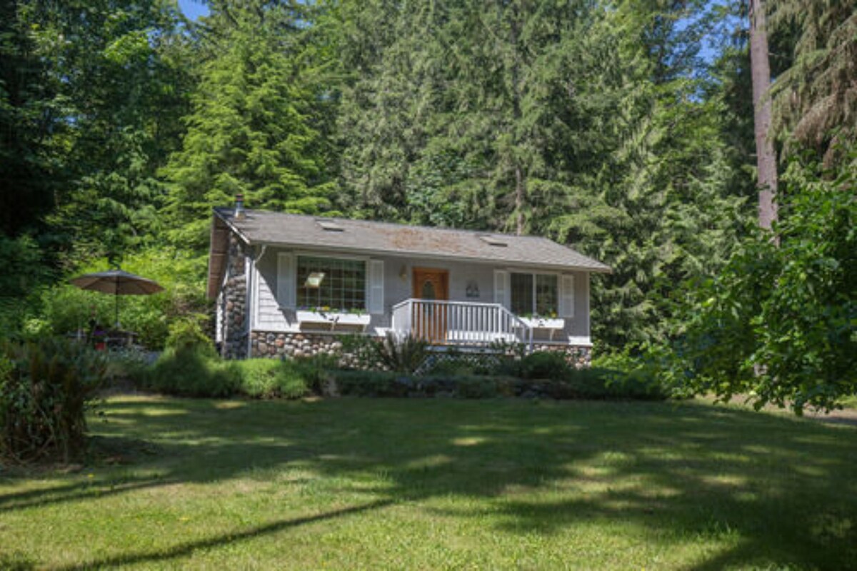 A single-story house with a front porch, surrounded by a well-maintained lawn and dense forest.