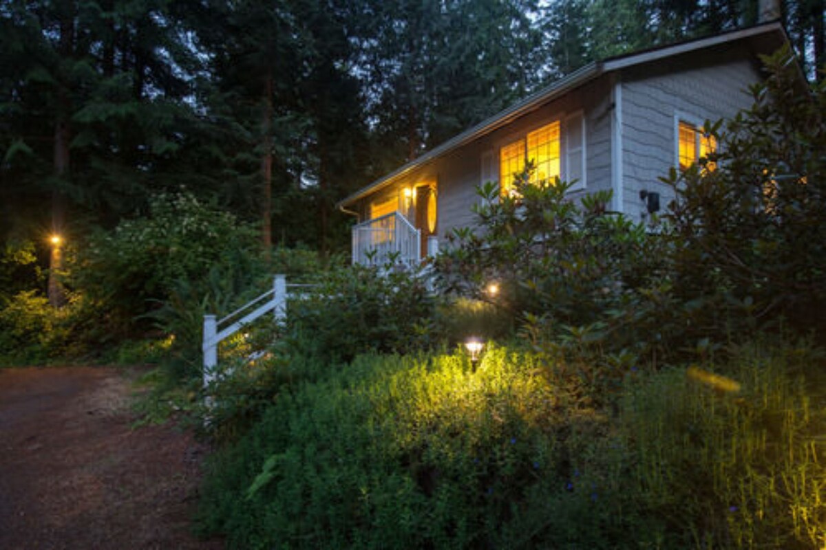 A house surrounded by dense vegetation with a white gate and pathway leading to the entrance.