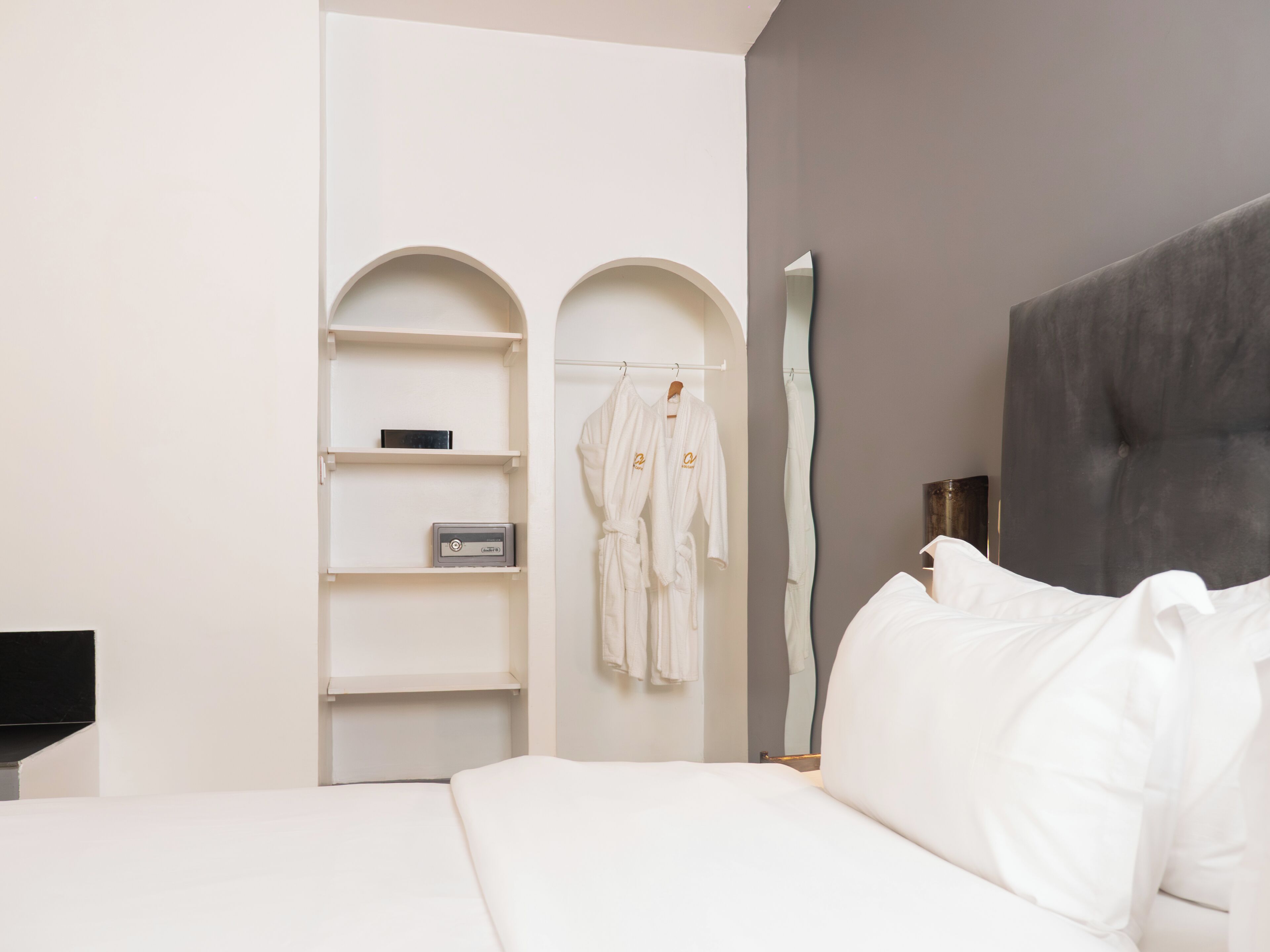 A bedroom with a bed, a wardrobe with arched shelves, and two white robes hanging on a rack.