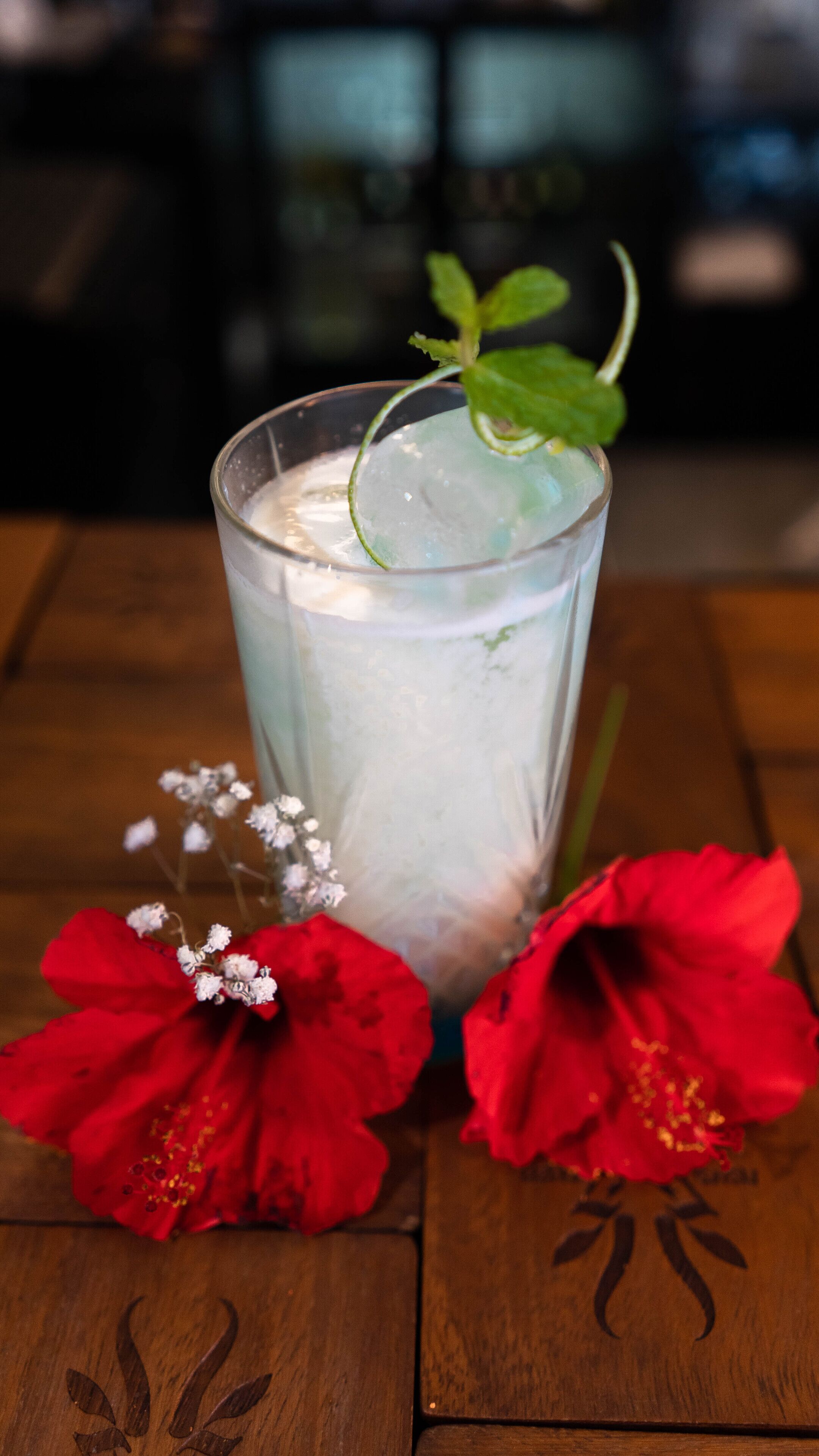 Een glas ijskoud drankje met een muntblaadje, samen met rode bloemen op een houten tafel.