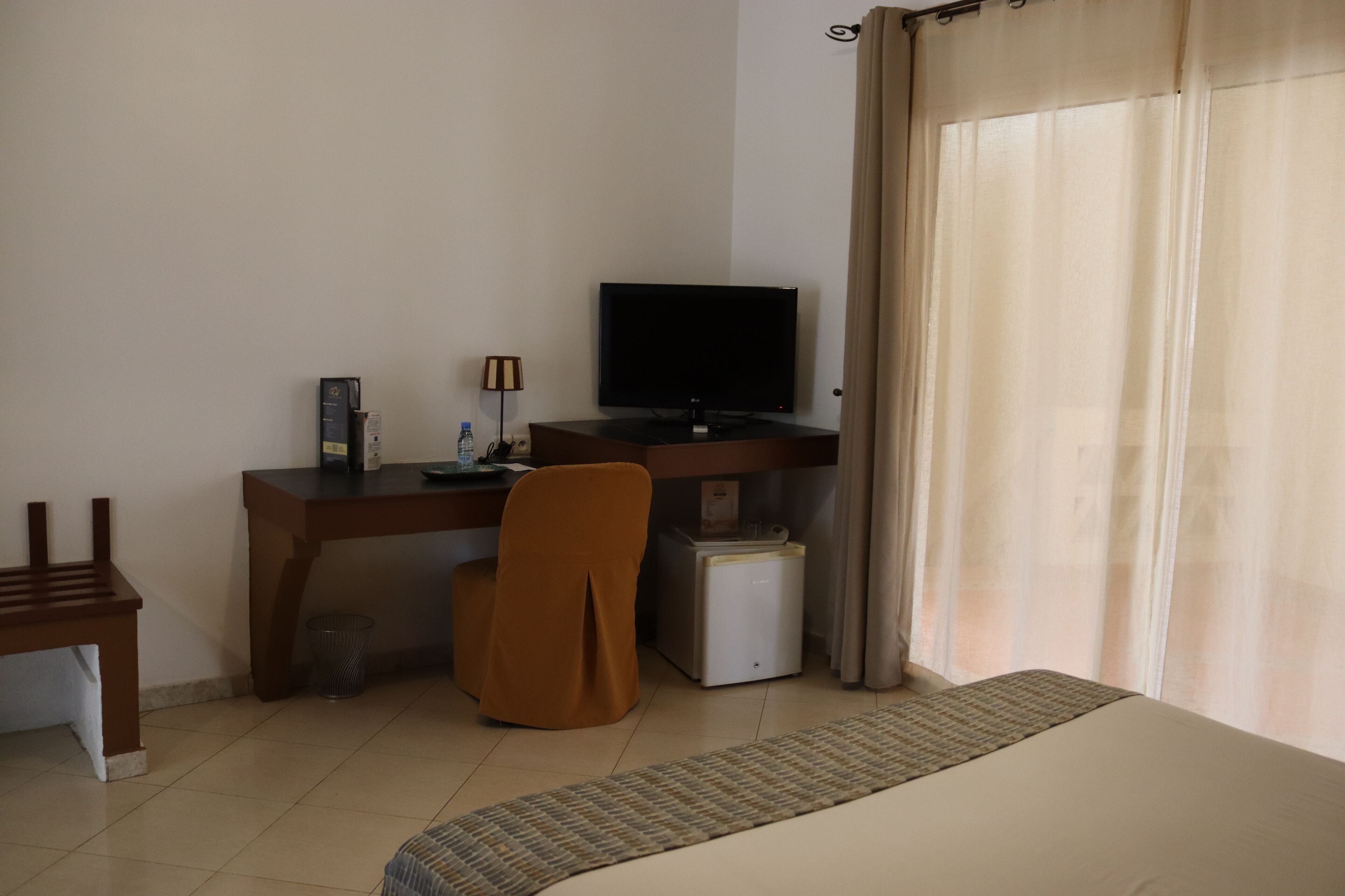 A hotel room with a bed, a desk with a computer, a chair, a small refrigerator, and a television.