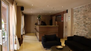 Un vestíbulo de hotel con un mostrador de recepción, dos sofás negros, una mesa de centro redonda y un ventanal con cortinas.