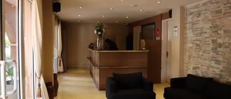 A hotel lobby with a reception desk, two black sofas, a round coffee table, and a large window with curtains.