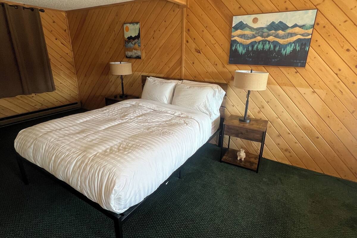 A hotel room with a wooden paneled wall, a bed with white bedding, two bedside tables with lamps, and a painting on the wall.