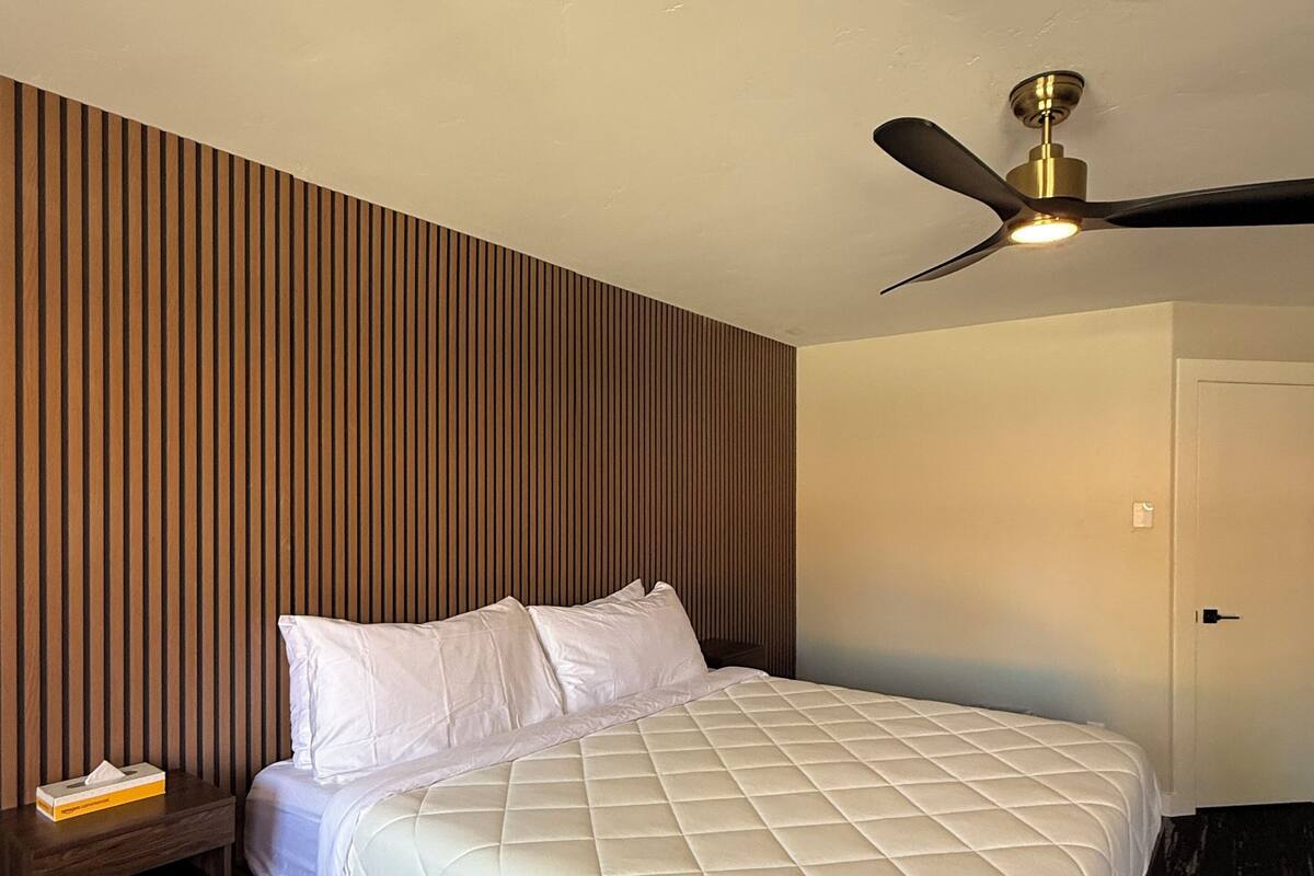 A hotel room with a bed, a nightstand, a wooden slat wall, a ceiling fan, and a door.