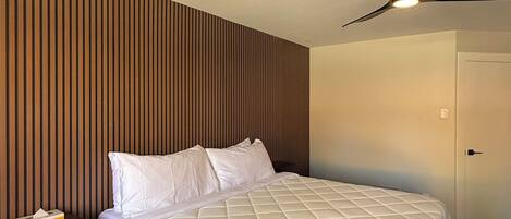 A hotel room with a bed, a nightstand, a wooden slat wall, a ceiling fan, and a door.