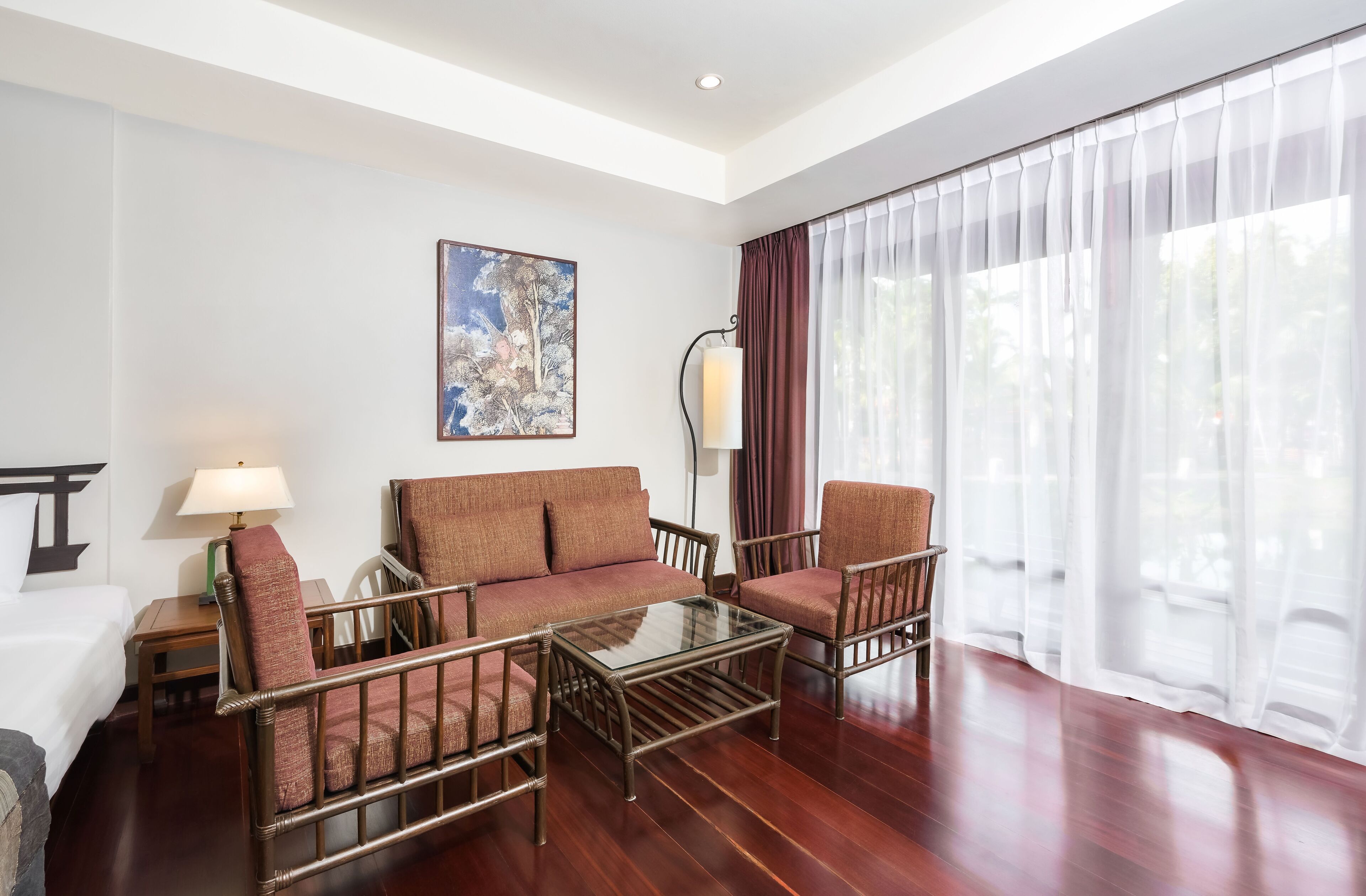 A hotel room with a sofa set, a glass coffee table, a lamp, and a painting on the wall.