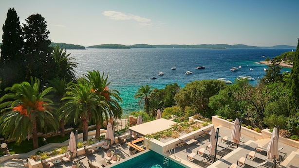 A poolside area with lounge chairs and umbrellas, overlooking a clear blue sea with boats.