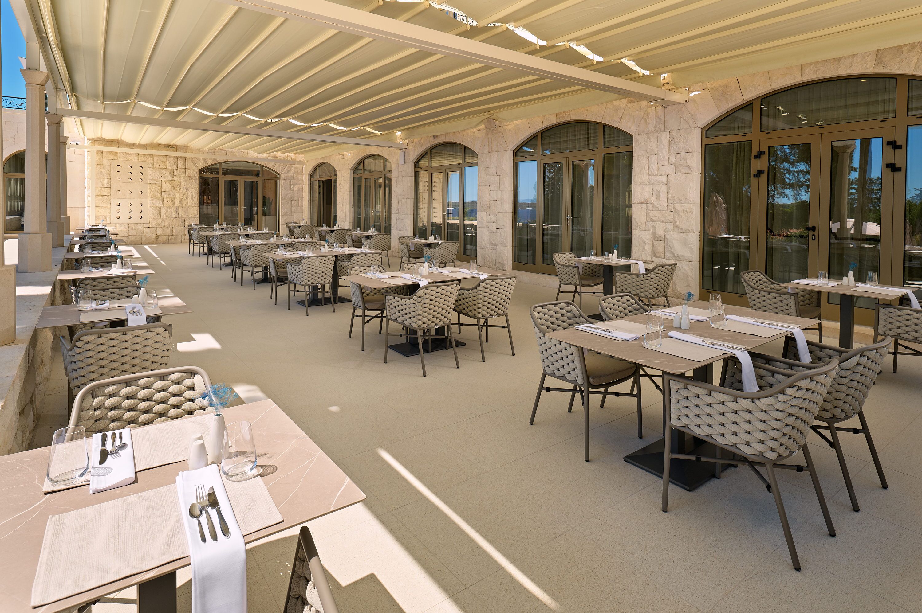 Un área de comedor al aire libre con varias mesas y sillas, paredes de piedra y ventanas grandes.