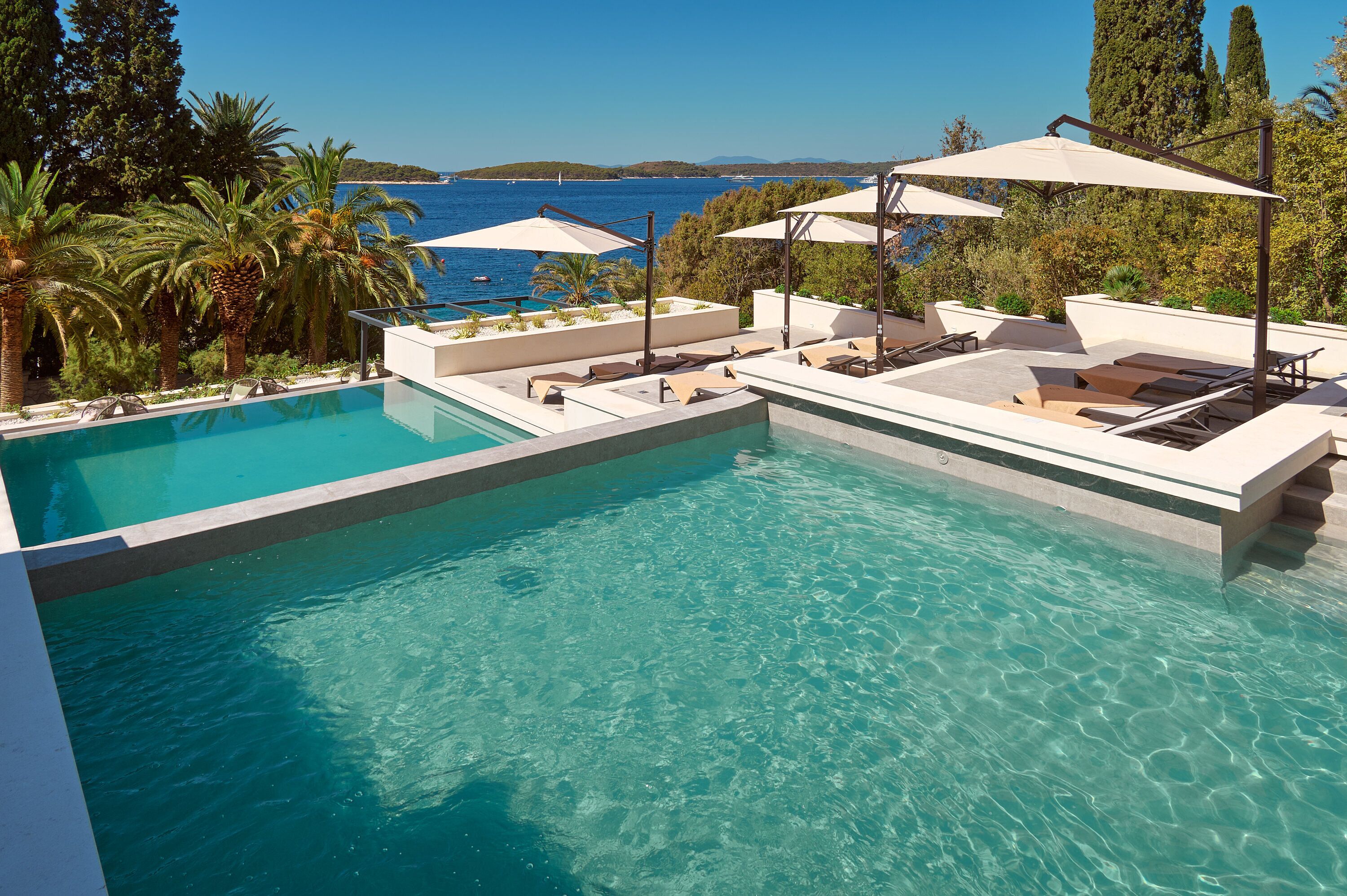 Une piscine d’eau cristalline, bordée de chaises longues et d’ombrelles, entourée de verdure et offrant une vue sur la mer.