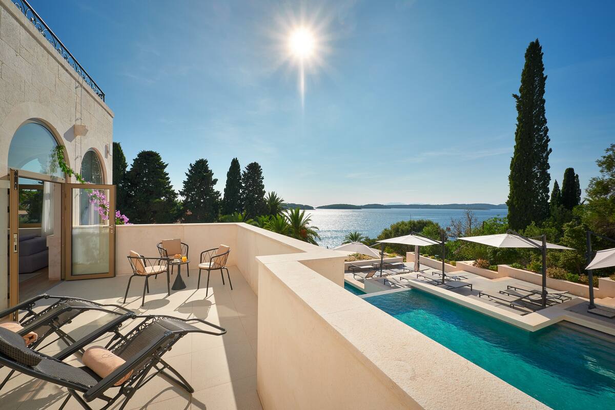 Un área junto a la piscina con sillones, una mesa y sombrillas, con vista a una masa de agua y un cielo despejado.