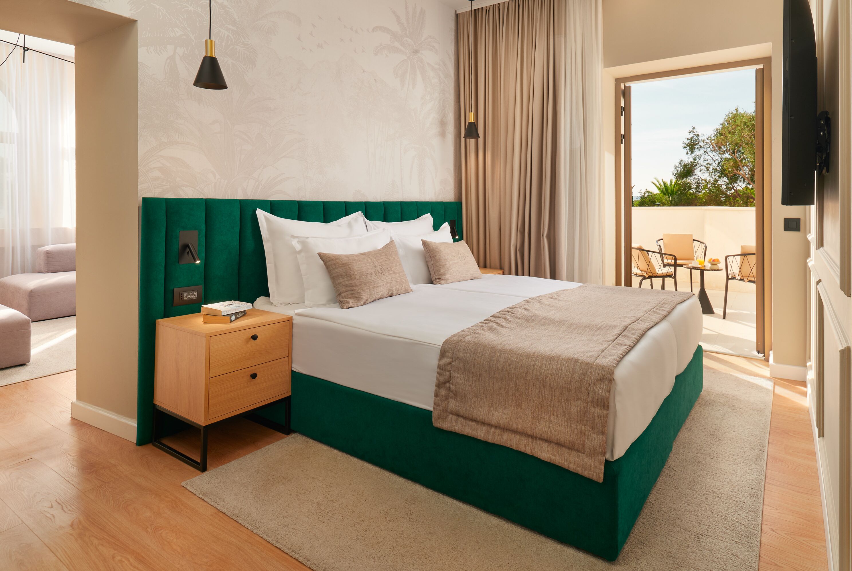 A hotel room with a green upholstered headboard, a large bed with white linens, a wooden nightstand, a beige rug, and a view of the outdoor patio area.