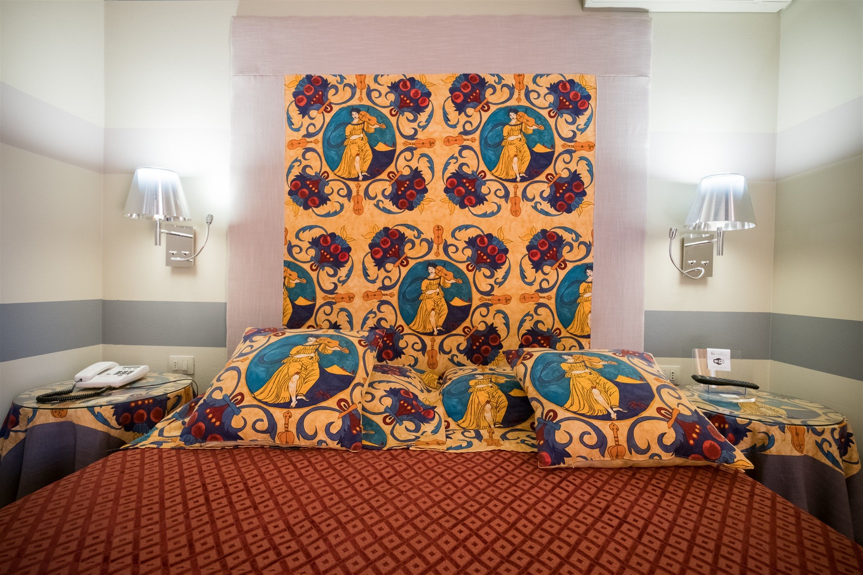 A hotel room with a patterned headboard, two bedside tables with lamps, a telephone, and a bed with a patterned bedspread.