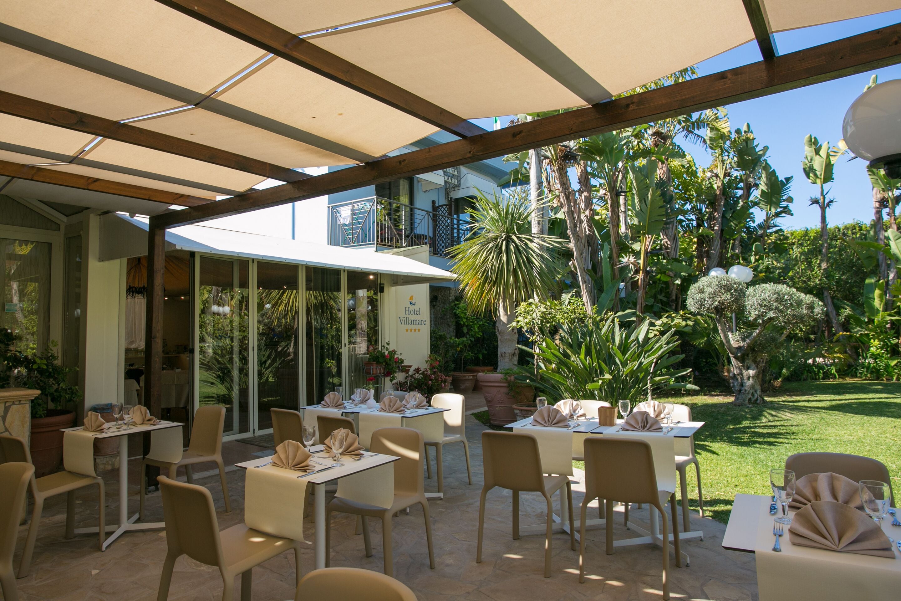 An outdoor dining area with tables and chairs, surrounded by lush greenery and a clear sky.