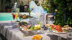 A table set with various foods and drinks, including a bottle of champagne, plates of appetizers, and glasses of water and juice, by a poolside. - Hotel Villamare (Syracuse)
