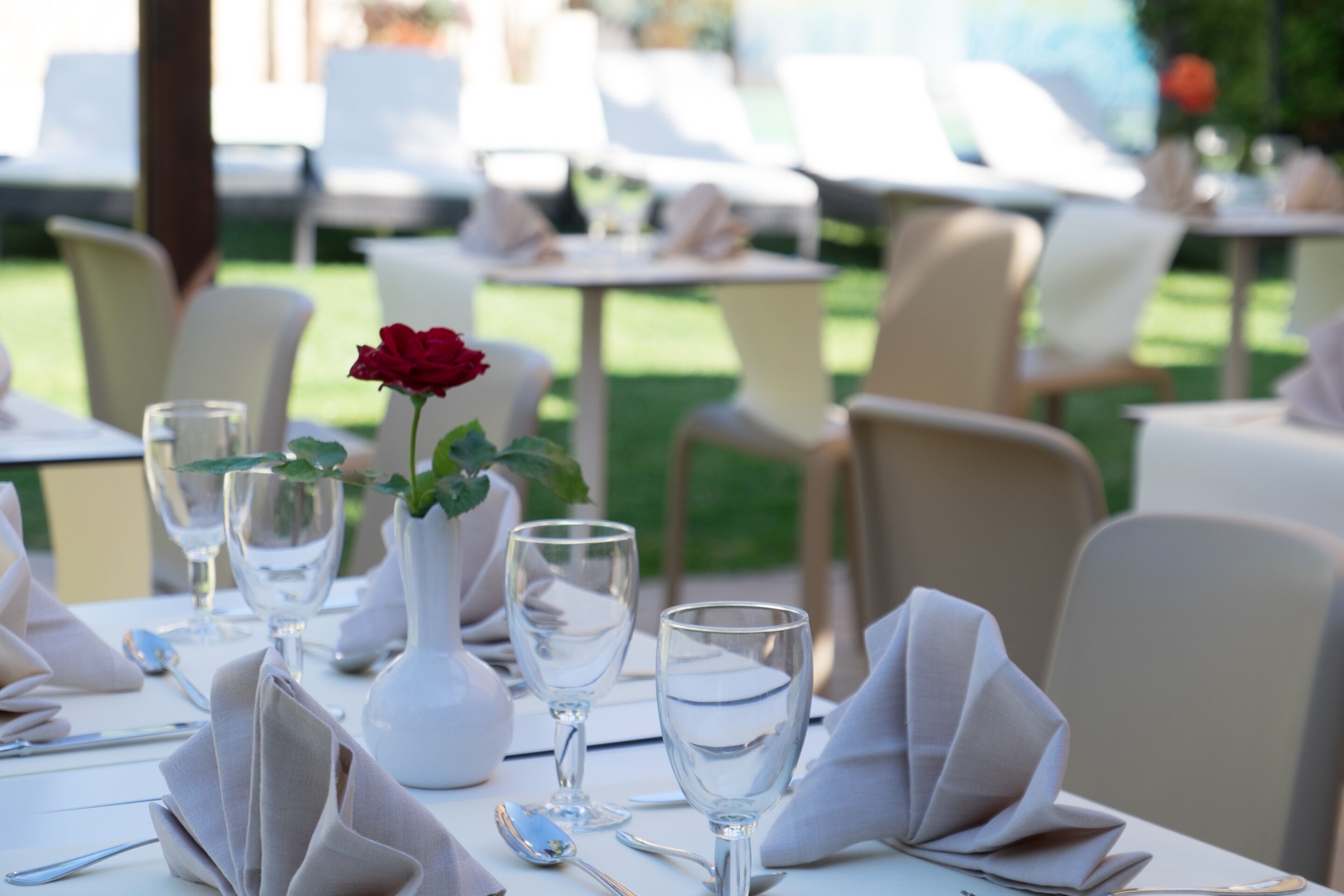 Une table à manger dressée avec des serviettes blanches, des verres à vin et un vase contenant une rose rouge.