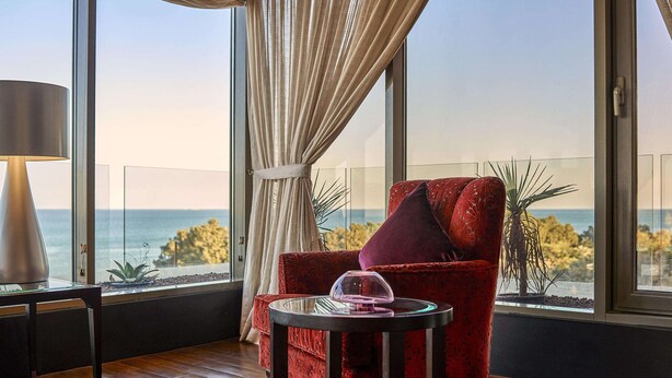 A room with a large window, a red chair, a small table, and a lamp. - Safir Fintas Kuwait Hotel (Fintas)