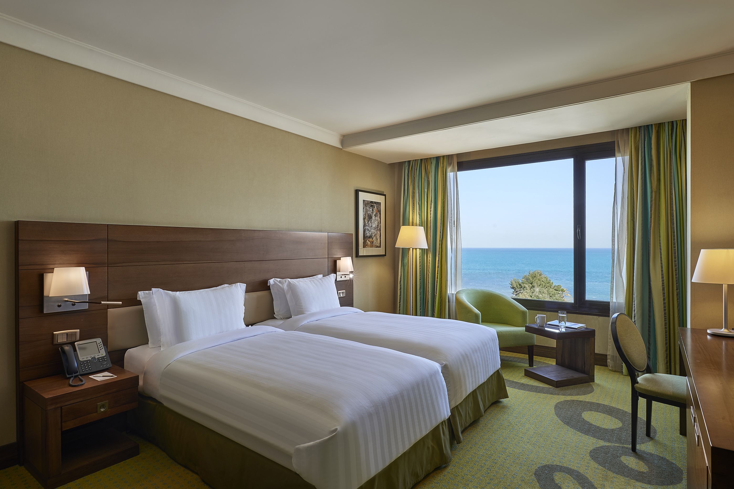A hotel room with two beds, a desk, a chair, a phone, and a window with a view of the sea.