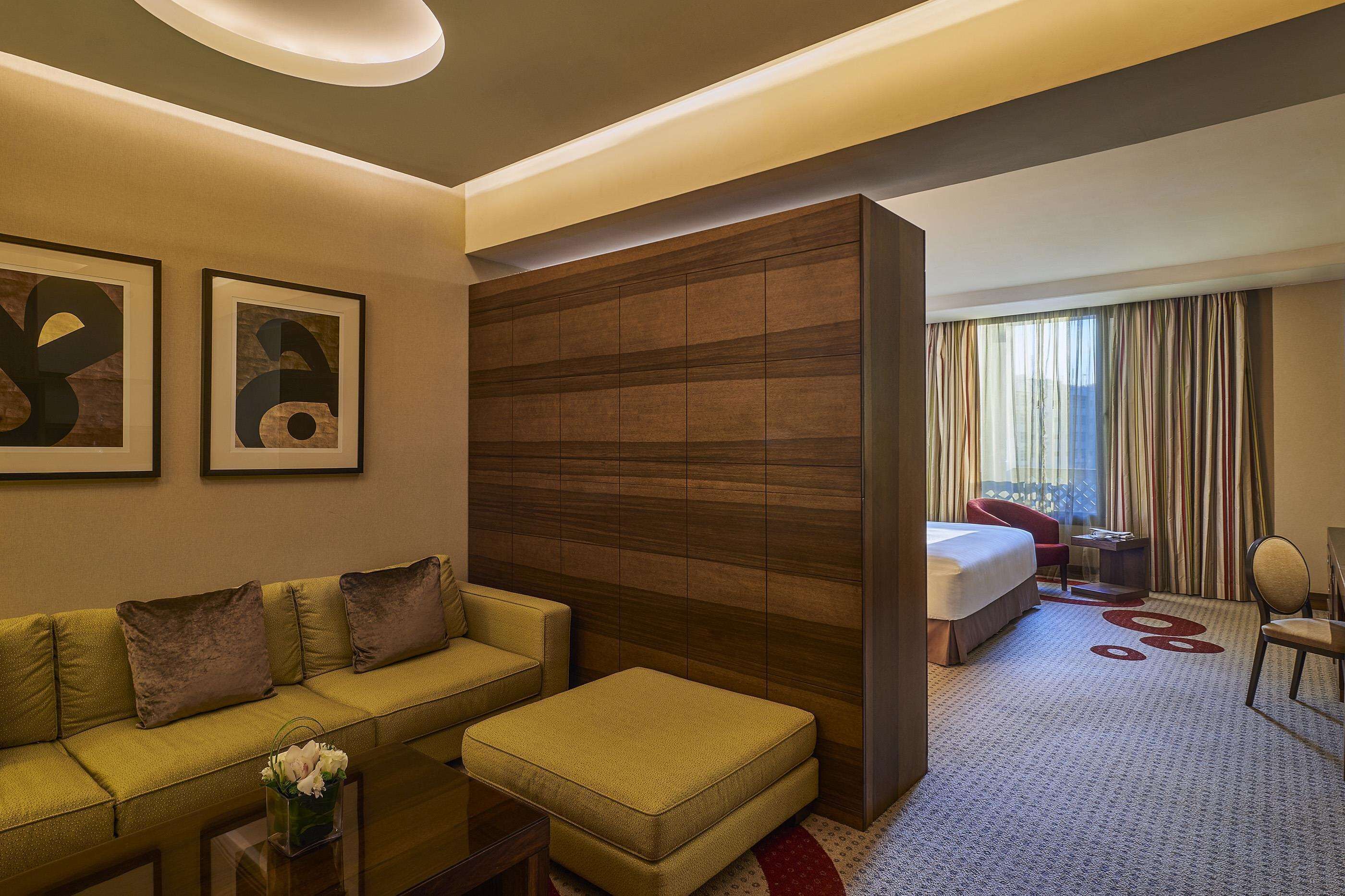 A hotel room with a sofa, ottoman, a bed, a small table with a vase of flowers, and a large wooden panel wall.