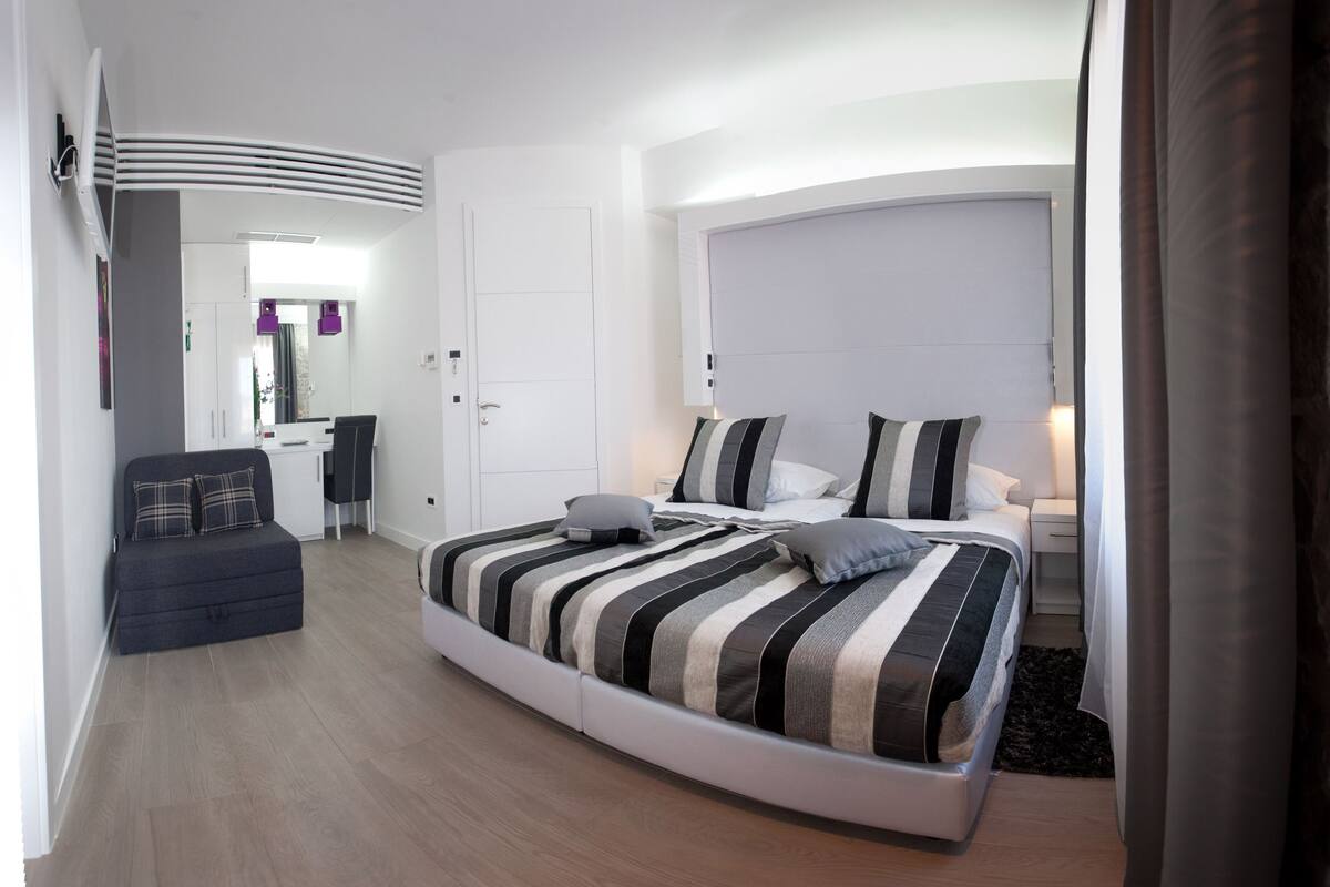 A modern bedroom with a curved bed, a small seating area, and a bathroom visible in the background.
