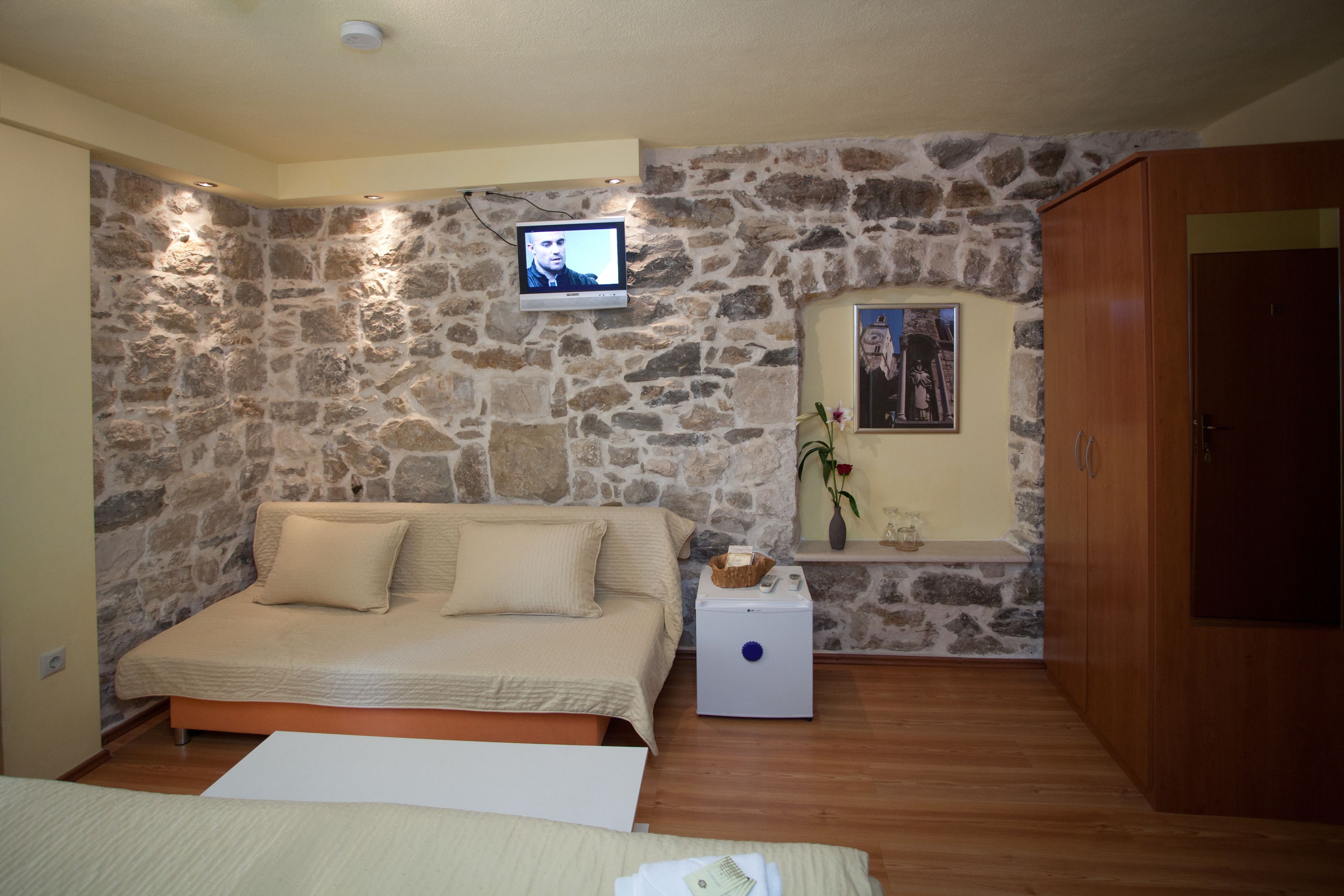 A bedroom with a stone wall, a bed, a small white table, a wooden wardrobe, and a television mounted on the wall.
