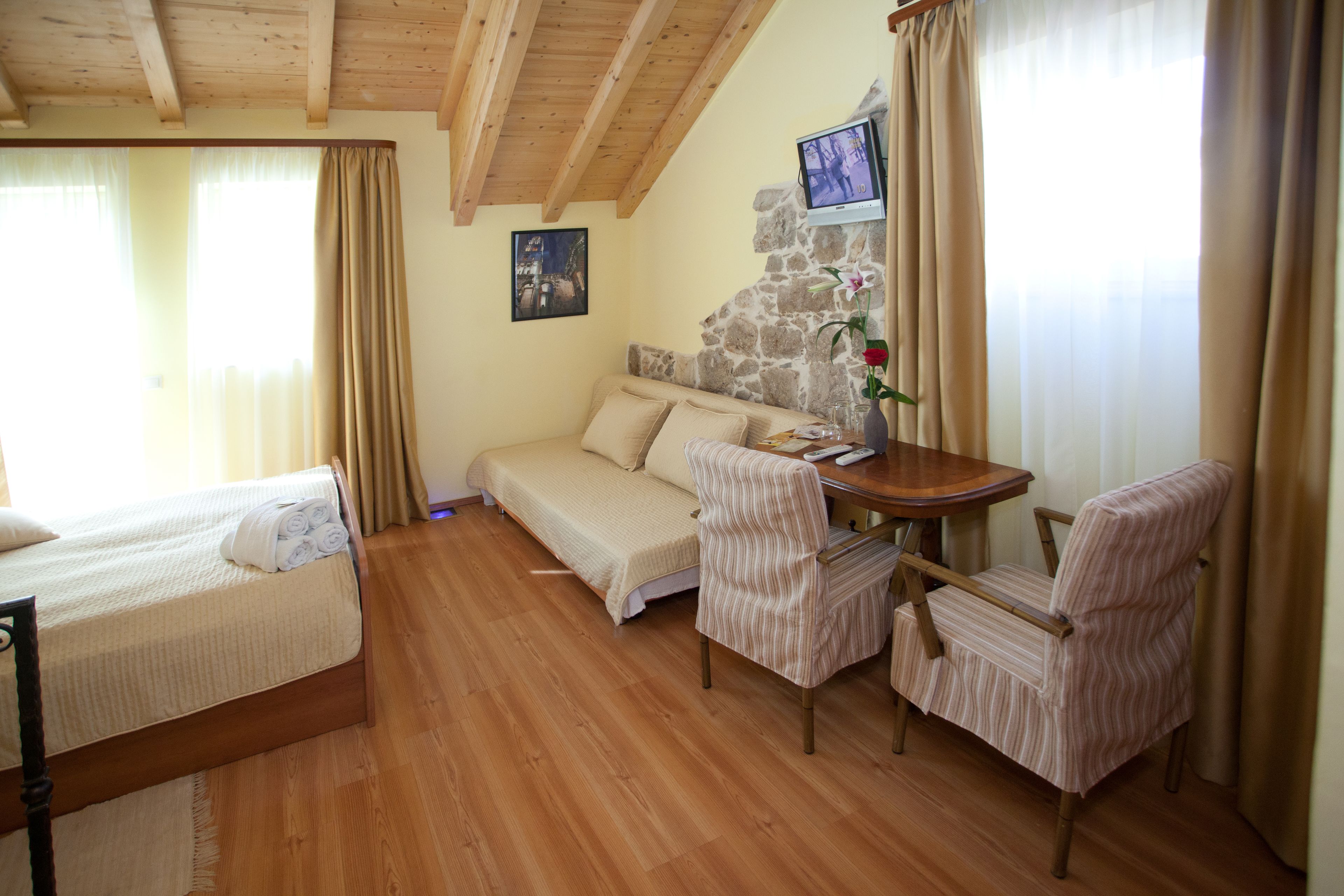 A room with a bed, a sofa, a dining table with chairs, a stone wall, and a TV mounted on the wall.