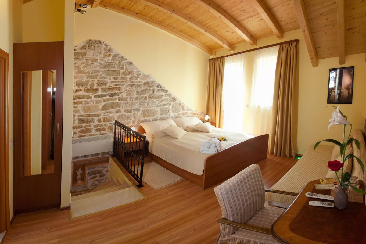 A bedroom with a stone wall, wooden ceiling, a bed with white linens, a chair, a small table with a vase of flowers, and a remote control.