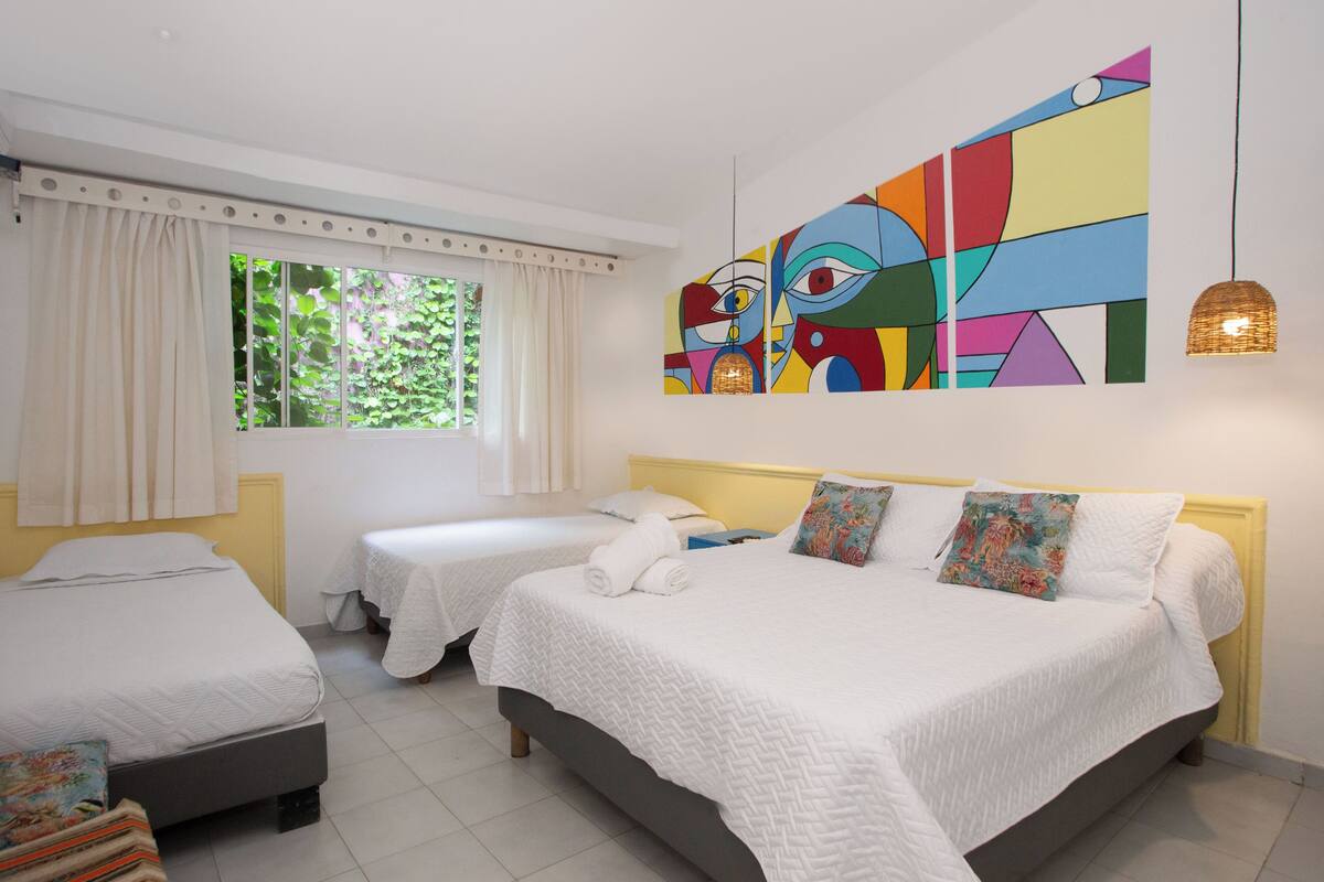 Et hotelværelse med to senge, et stort vindue med gardiner, et farverigt abstrakt maleri og en hængende lampe.