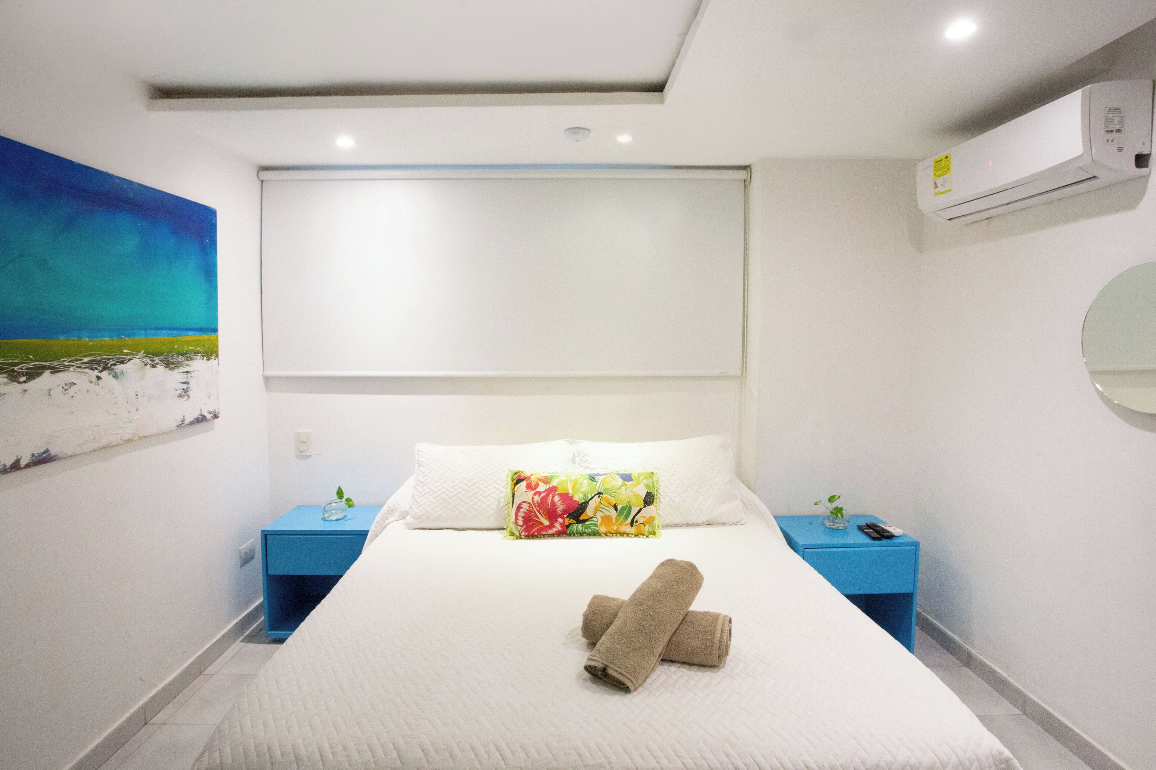 A hotel room with a large bed, a painting on the wall, a blue nightstand, and an air conditioning unit.