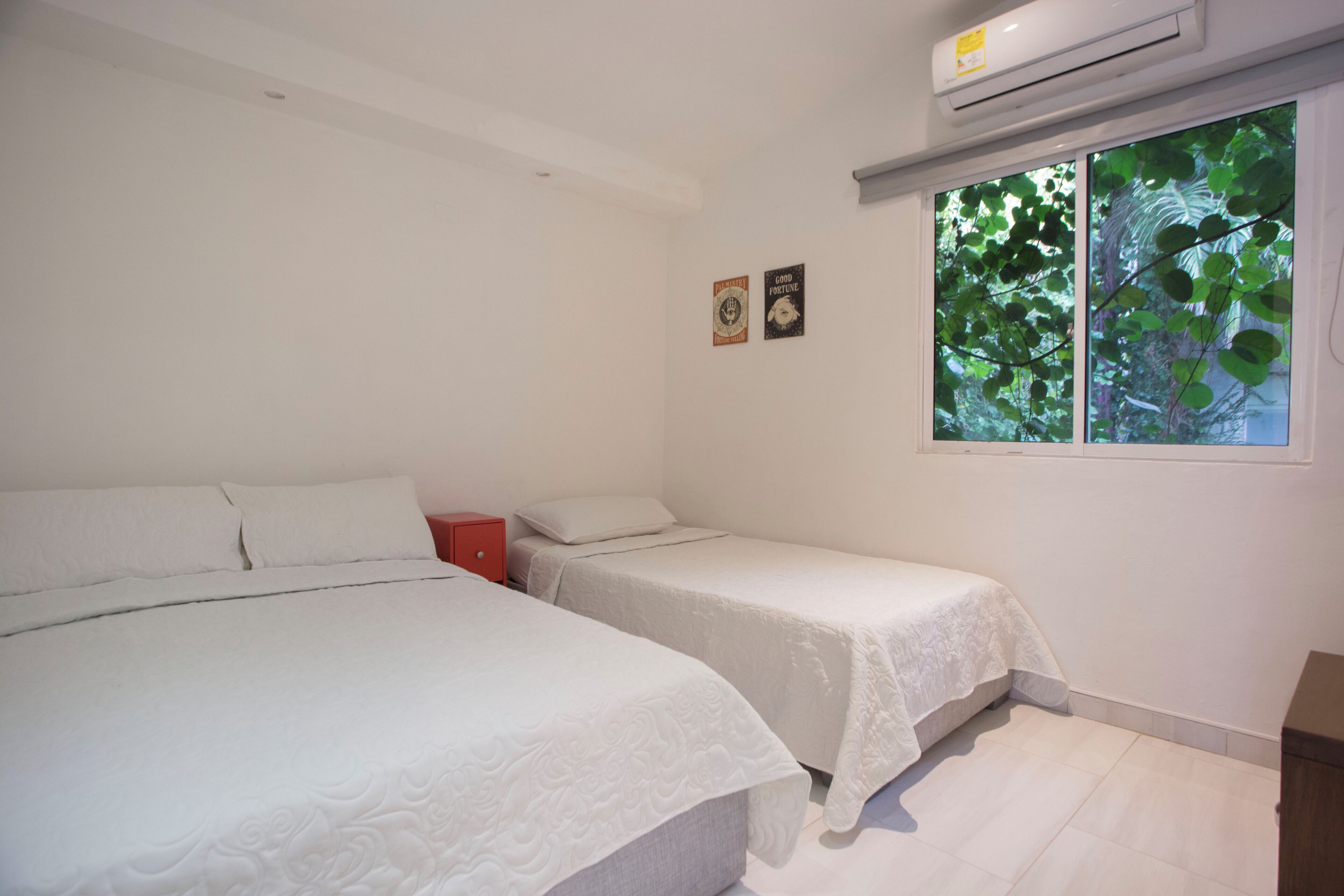 A bedroom with two single beds, a red nightstand, and a window with a view of trees.