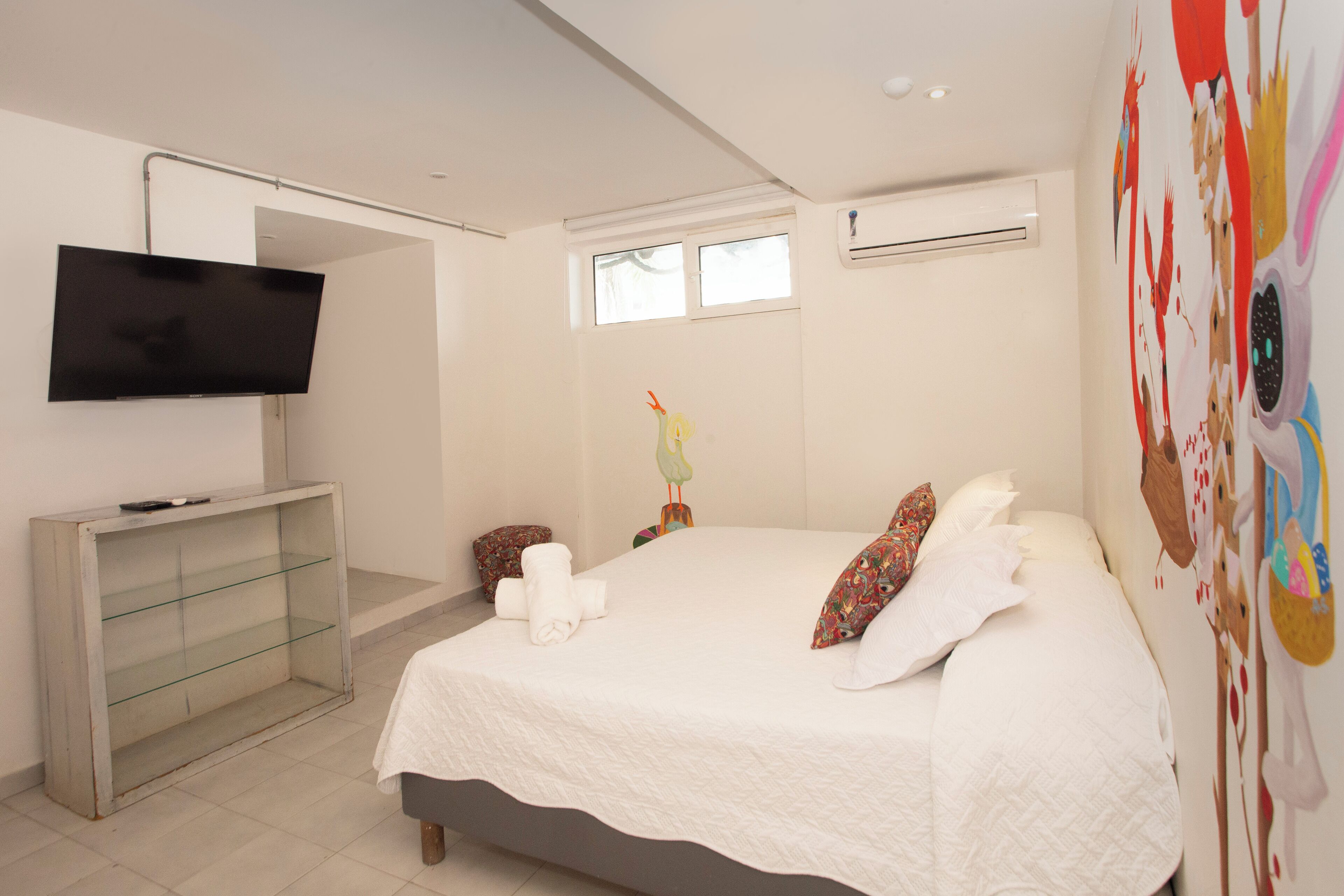 Quarto moderno com cama, televisão fixada na parede, pequeno estante e janela.