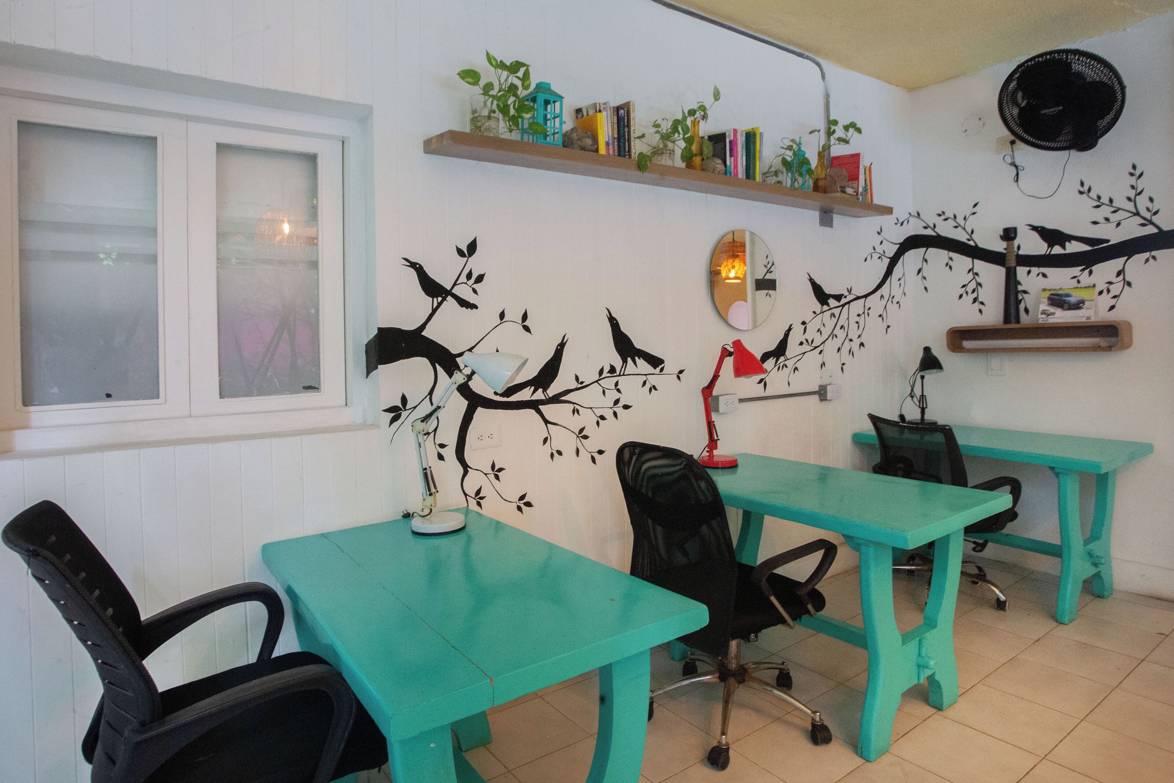A room with teal-colored tables and chairs, a white wall with a tree mural, a window with frosted glass, and a shelf with books and plants.