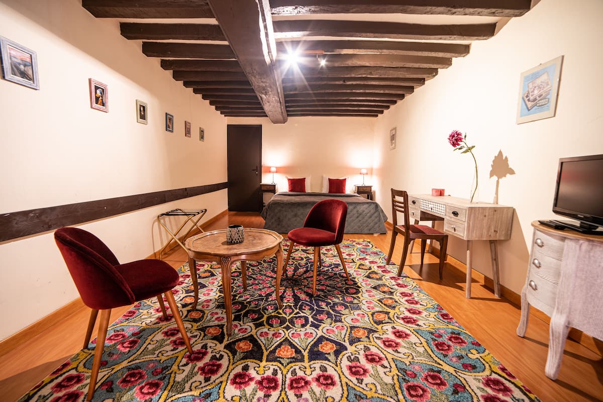 Una habitación acogedora con una cama, dos sillas rojas, una mesita, un televisor y una alfombra estampada.