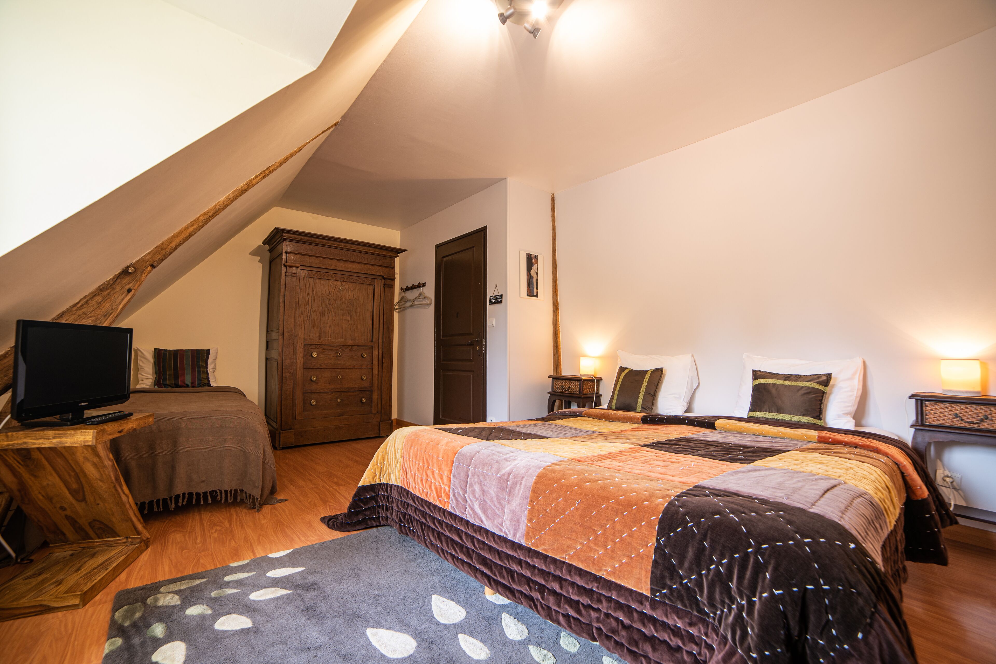 Un dormitorio con techo estilo loft, una cama grande con colcha, un aparador de madera, un televisor sobre una mesita auxiliar y una mesita de noche con lámpara.