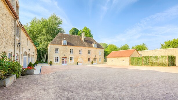 Une cour avec un grand bâtiment en pierre, un bâtiment plus petit en briques et une haie.