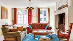Un salon avec un canapé beige, une table basse en bois et un fauteuil à rayures. On y trouve une cheminée avec une manteau de cheminée et une fenêtre avec des rideaux.
