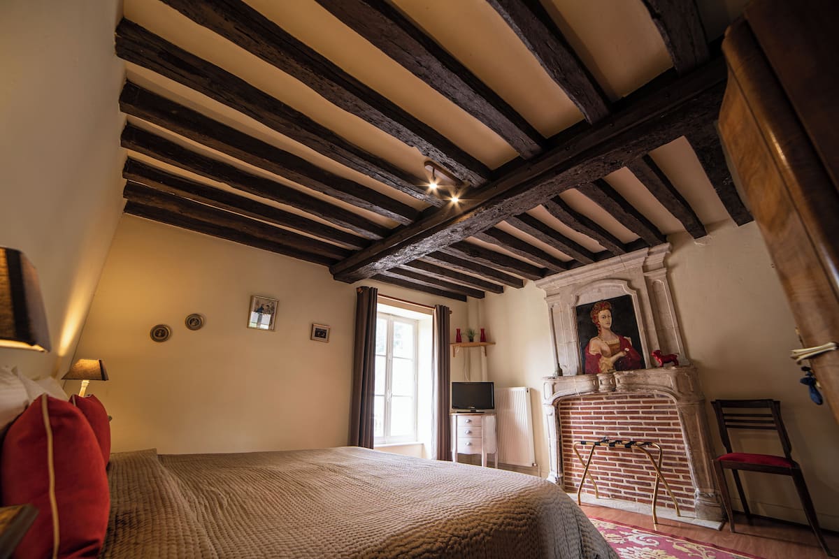 Un dormitorio con vigas de madera a la vista, chimenea, televisor y cama con almohada roja.