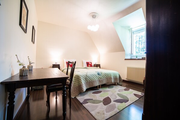 Un dormitorio con una cama, un escritorio y una silla. Hay una ventana con vistas a árboles y un radiador en la pared.