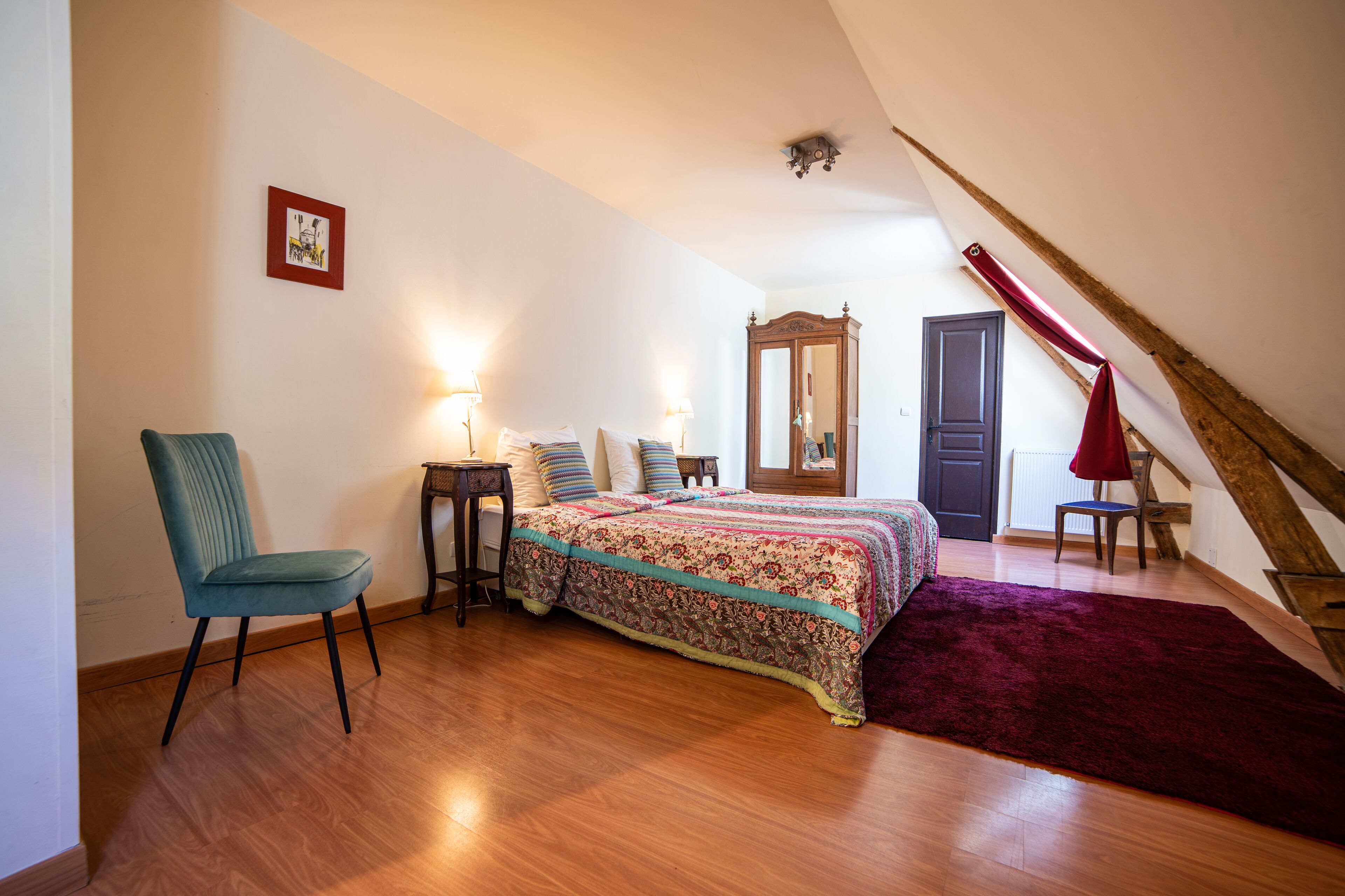 Un dormitorio con piso de madera, una cama con cubrecama estampado, una silla, una mesita de noche, un espejo y una cortina roja.