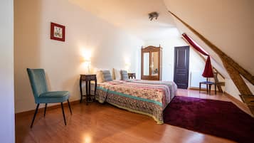 Un dormitorio con piso de madera, una cama con cubrecama estampado, una silla, una mesita de noche, un espejo y una cortina roja.
