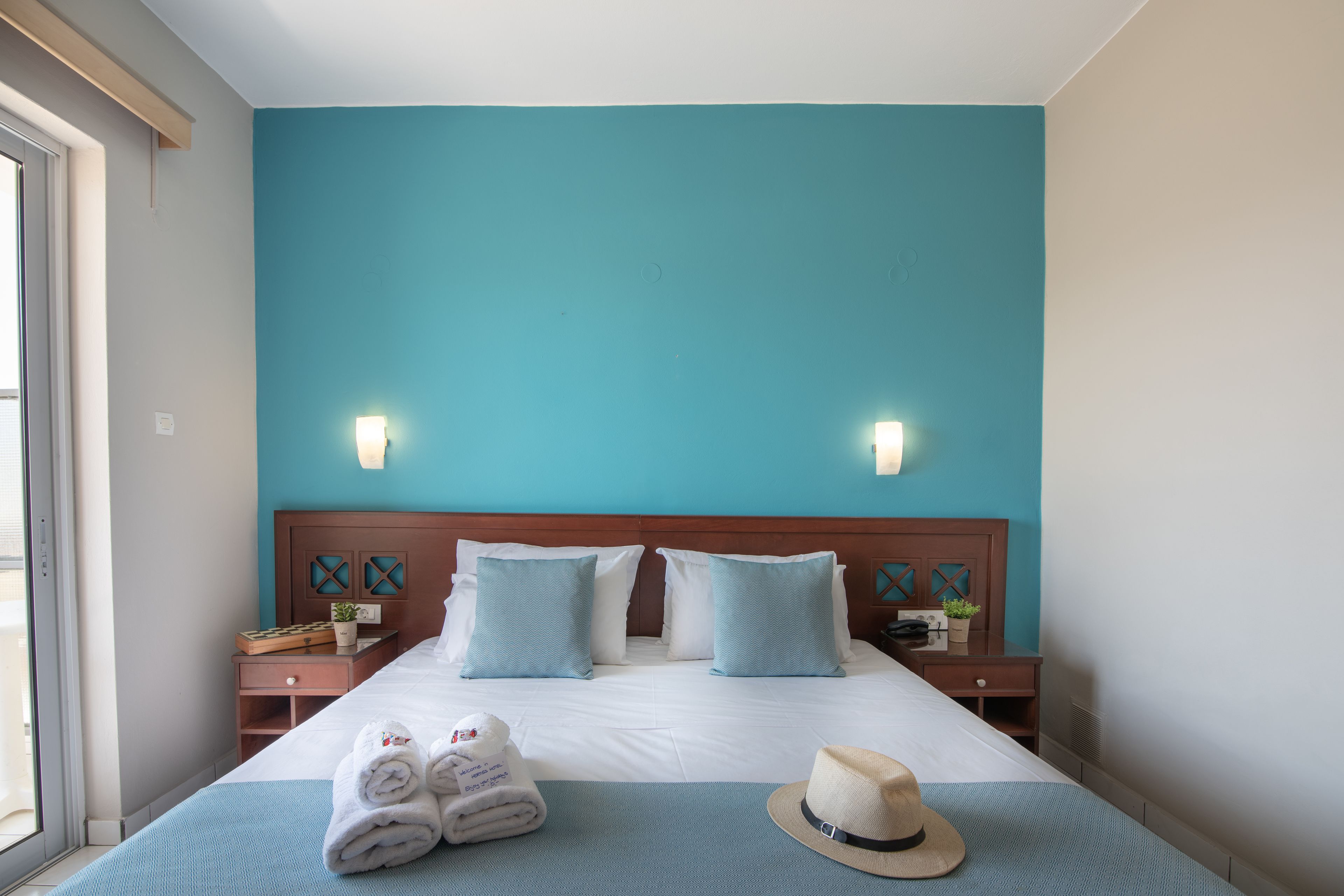 A hotel room with a large bed, two nightstands, a window, and a teal wall.