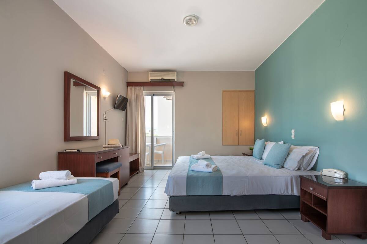 Habitación de hotel con una cama grande, una cama más pequeña, un escritorio, un espejo, un armario y una puerta corrediza de vidrio al balcón.
