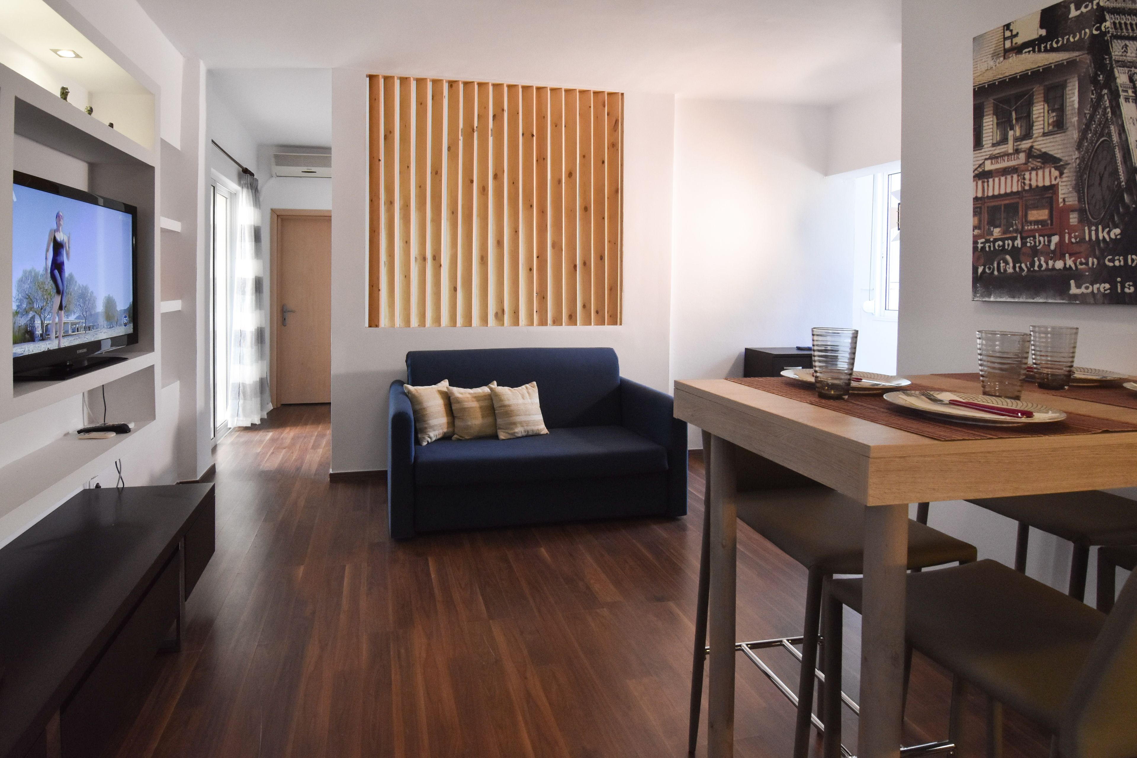 Un soggiorno moderno con un divano blu scuro, pannelli a listelli in legno, una televisione a schermo piatto e un'area pranzo con tavolo in legno e sgabelli da bar.